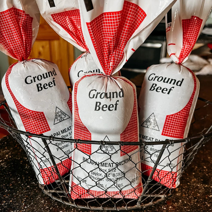 Four packages of Ground Beef, individually wrapped in white paper with red gingham patterns and tied at the top, arranged in a black wire basket on a dark countertop.
