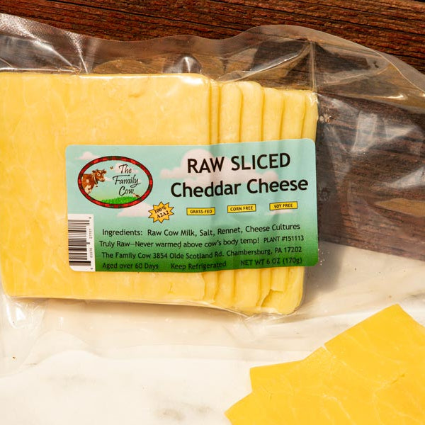 A package of raw sliced cheddar cheese in a clear plastic bag with a light blue and green label, shown on a white surface with a wooden background. Sliced cheese is also placed in the foreground.